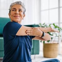 An older woman sitting on the floor while stretching one arm.